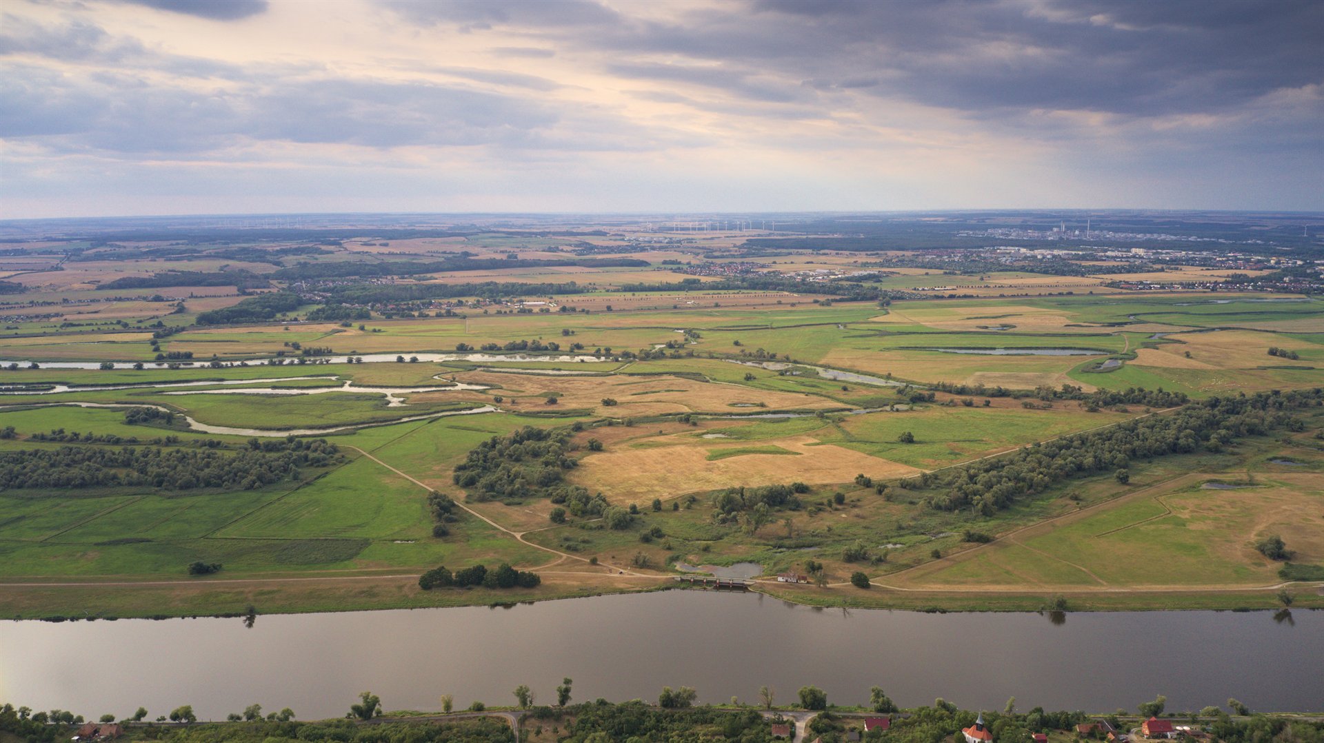Panorama wiejskiej doliny z meandrującą rzeką, zielonymi łąkami, polami i rozproszonymi zagajnikami przy pochmurnym niebie