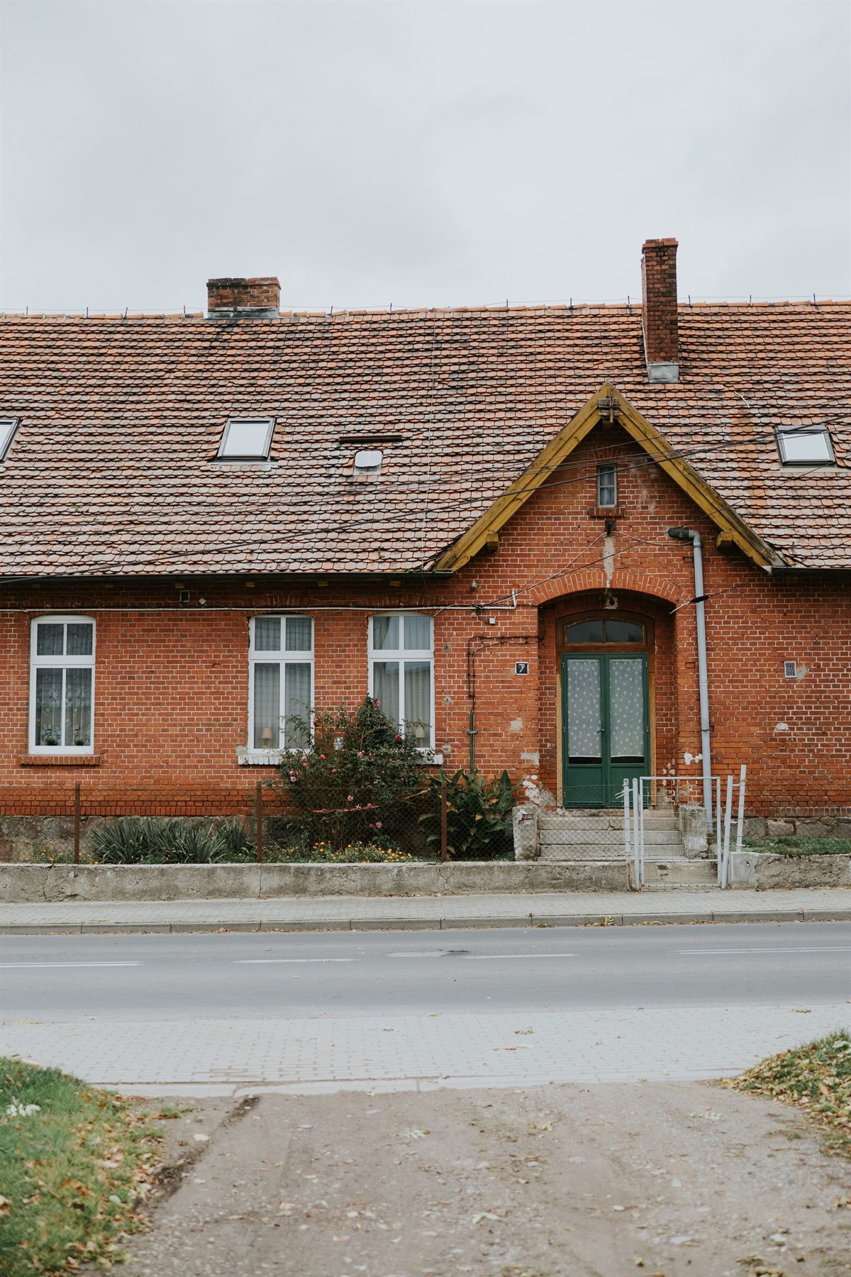 Czerwona ceglana kamienica z zielonym wejściem, dachem z dachówek i kwitnącymi krzewami przed oknami