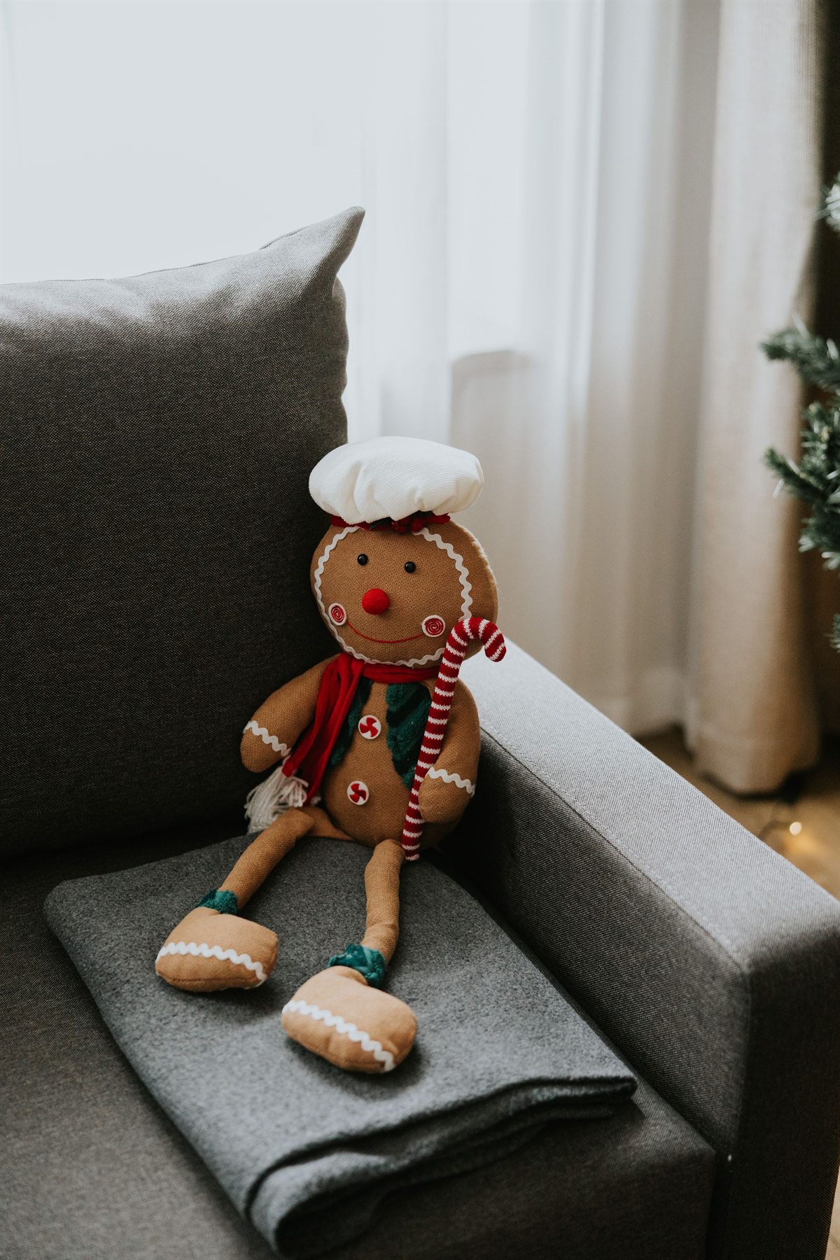 pluszowy piernikowy ludek z białą czapką, czerwoną szaliką i lizakiem siedzący na szarej kanapie
