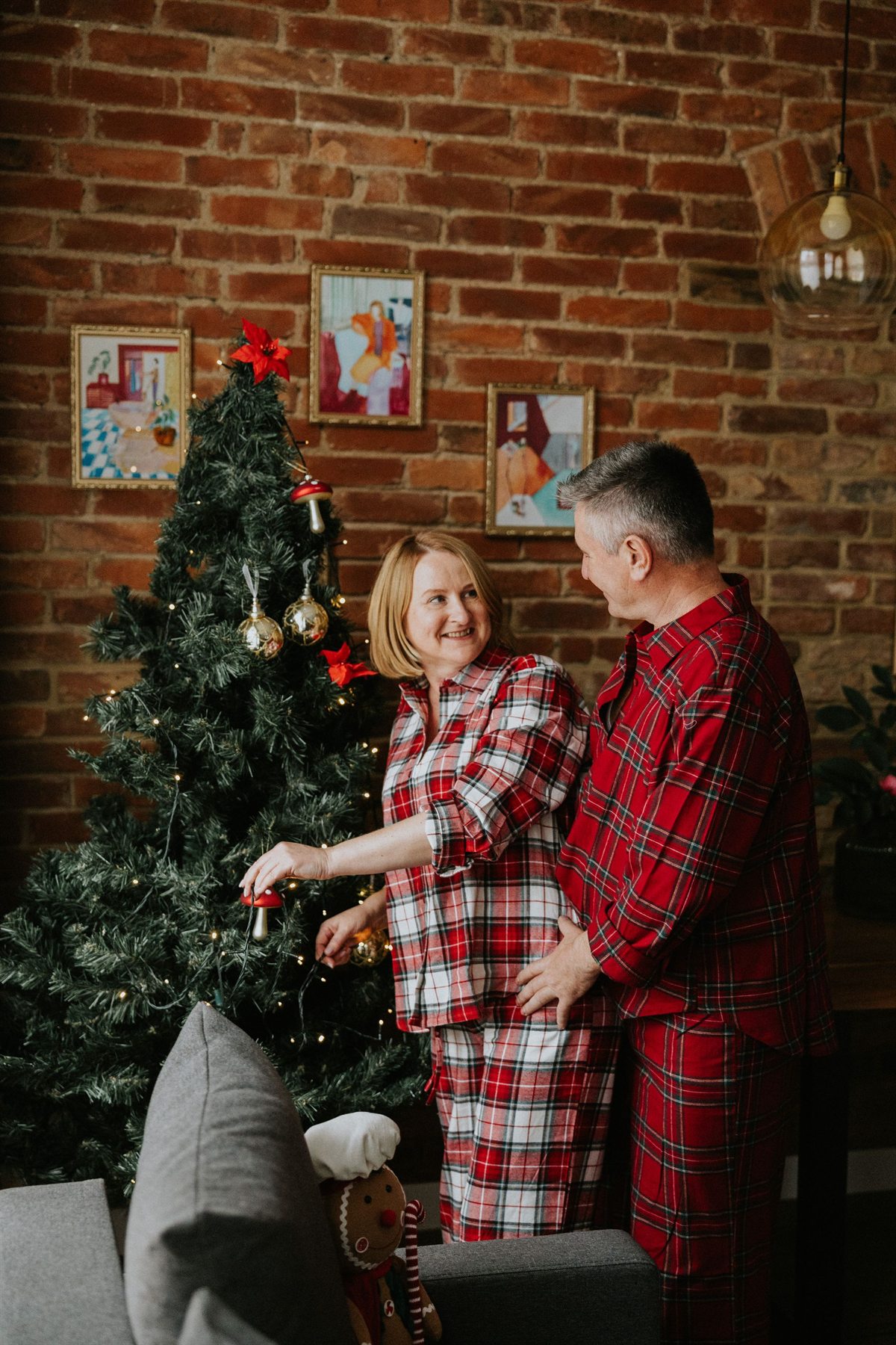 Para w czerwonych flanelowych piżamach ubierająca choinkę przy ceglanej ścianie, uśmiechnięci i trzymają ozdoby