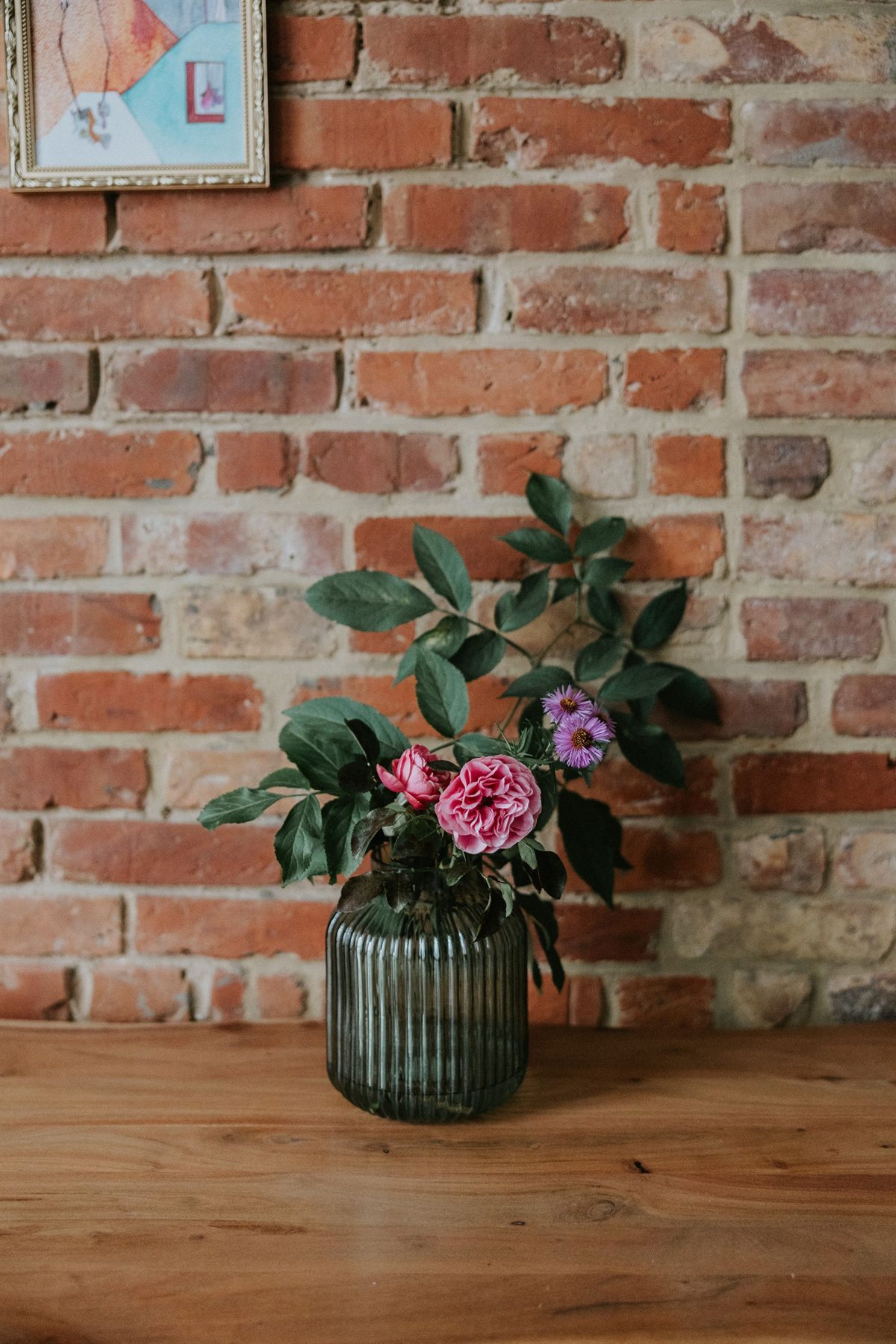 Szklany wazon z różowym kameliowym kwiatem, drobnymi fioletowymi astrami i zielonymi liśćmi na drewnianym stole przed ceglanym murem