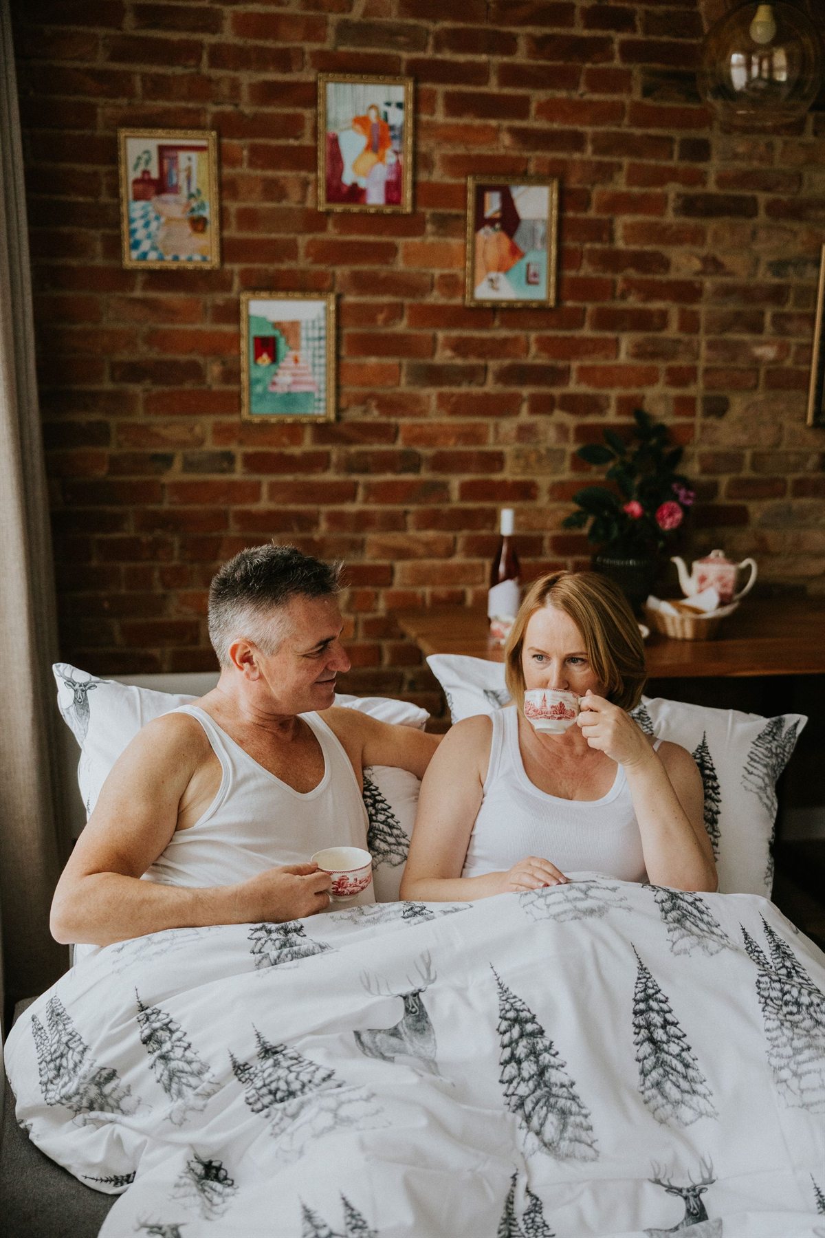 Para w białych podkoszulkach pijąca herbatę w łóżku z narzutą w choinki, ceglany mur i obrazki w tle