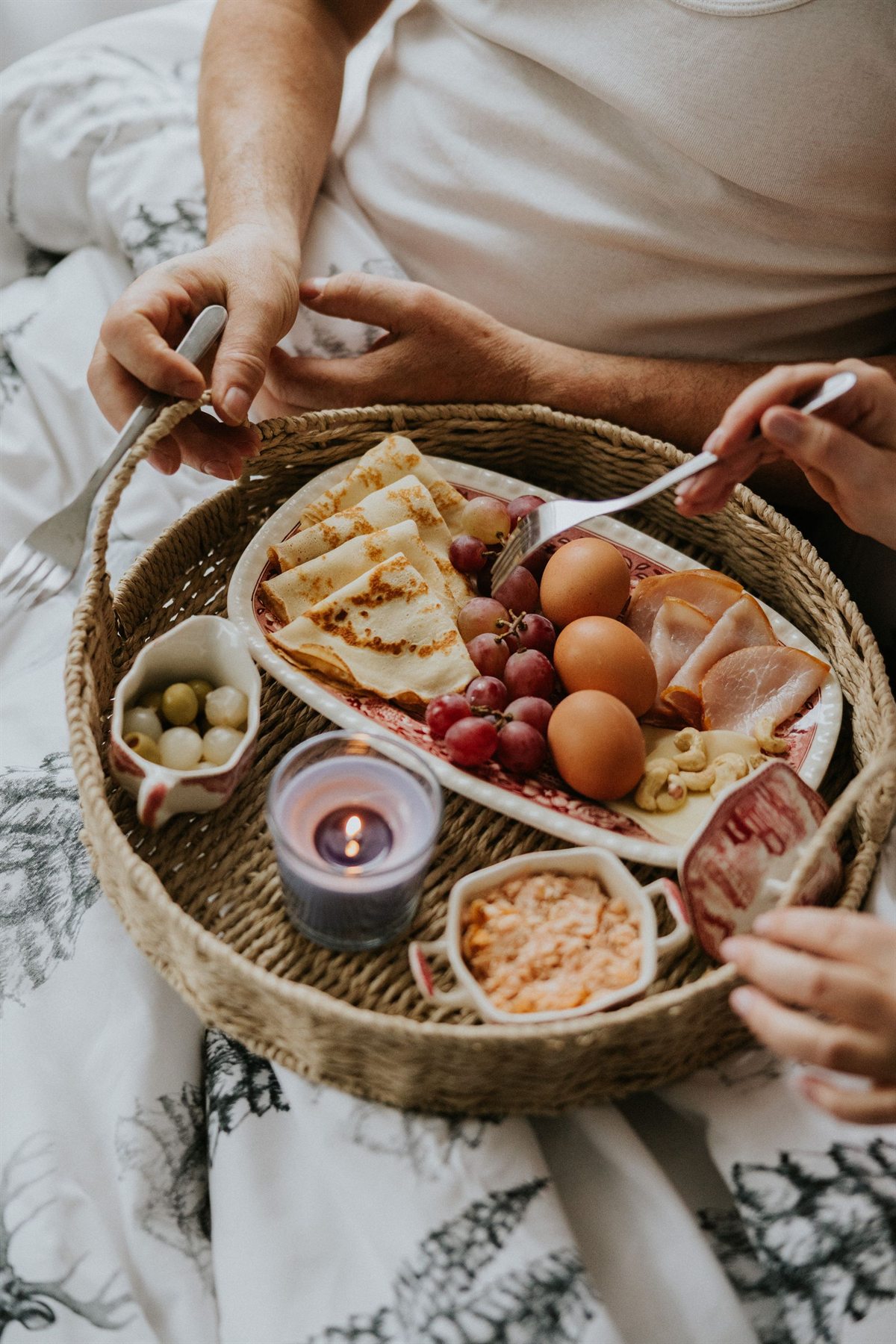 Taca śniadaniowa z naleśnikami, winogronami, jajkami, szynką, oliwkami i zapaloną świecą na łóżku