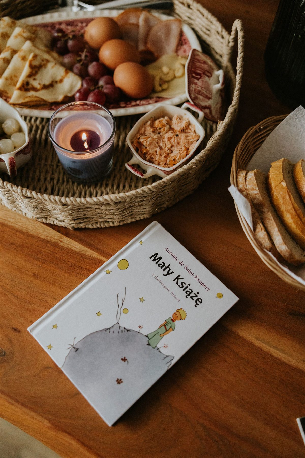 Okładka książki "Mały Książę" na drewnianym stole obok kosza z pieczywem, świecy i przekąsek
