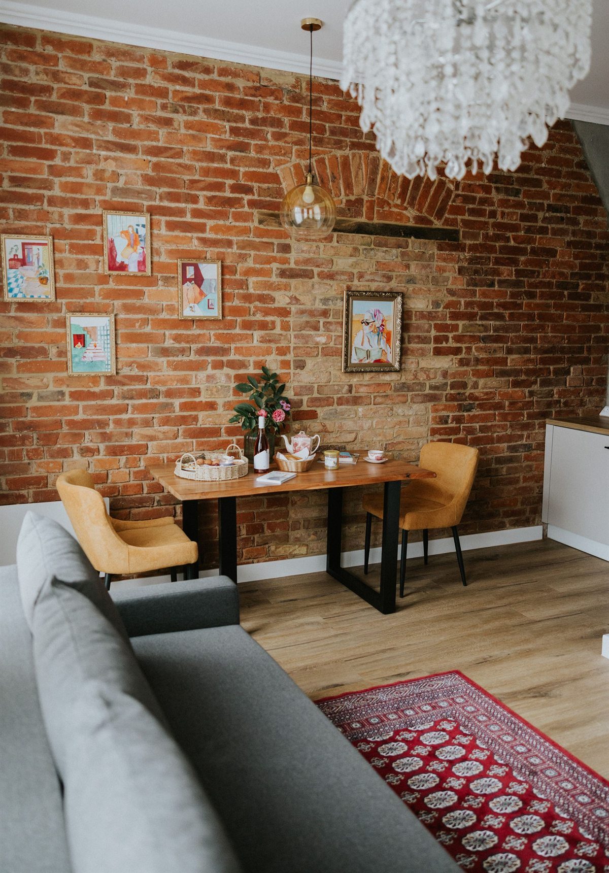 Przytulne wnętrze z ceglanym murem, drewnianym stołem, żółtymi krzesłami, kwiatami i szarą sofą