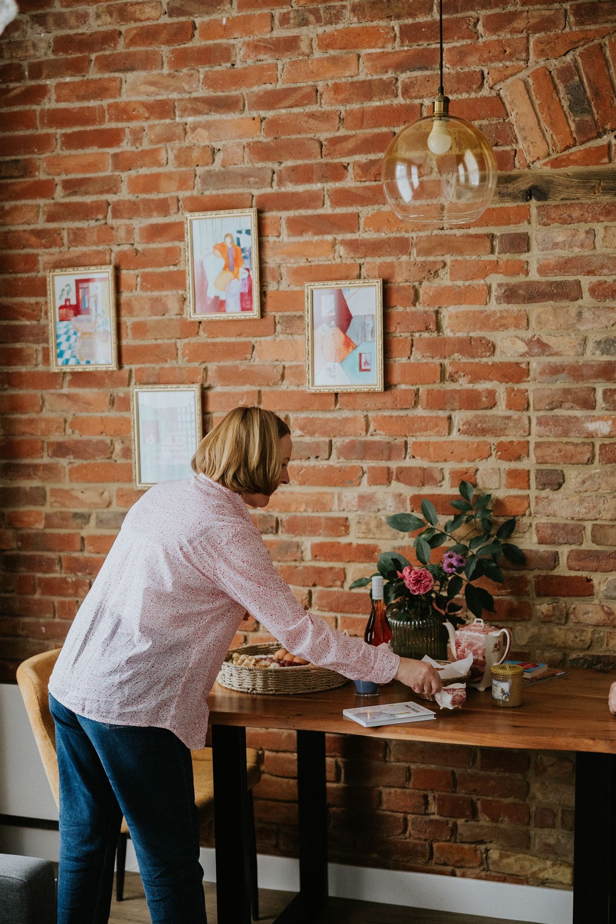 Kobieta ustawiająca filiżankę na drewnianym stole z wazonem kwiatów i koszem, ceglana ściana z obrazkami w tle