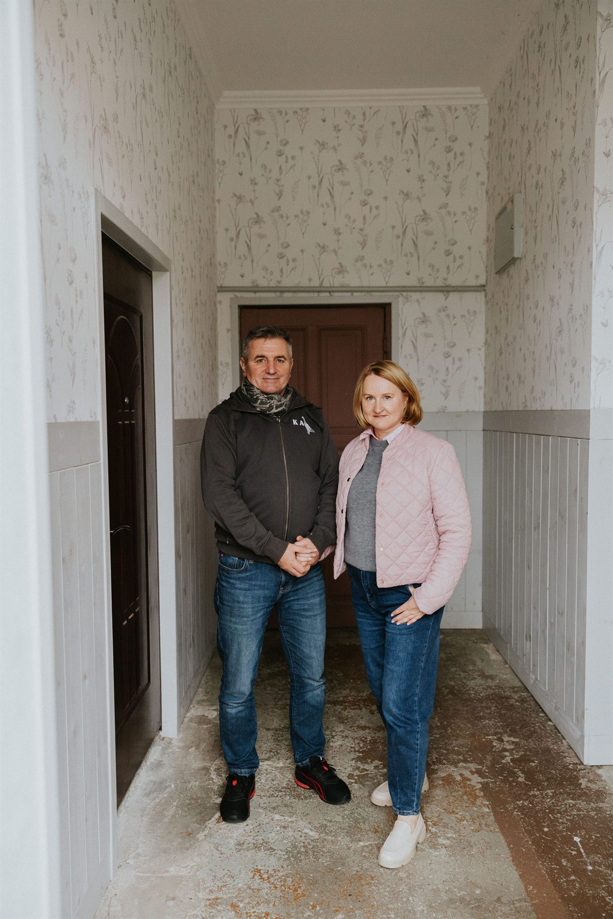 Para w średnim wieku stojąca w wąskim korytarzu; mężczyzna w ciemnej bluzie i kobieta w różowej pikowanej kurtce